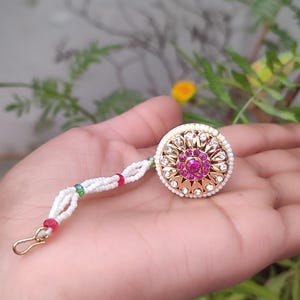 May include: A gold-coloured hair accessory featuring a central pink gemstone, surrounded by clear stones and a white beaded chain. The accessory has a circular design with a hook clasp.