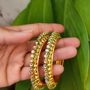 Peut inclure: Deux bracelets dorés ornés de pierres précieuses rondes et transparentes et d'accents verts. Les bracelets présentent un design détaillé et sont présentés sur un fond de feuilles vertes. Les bijoux sont ornés et accrocheurs.