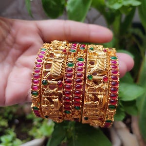 May include: A collection of gold-coloured bangles with detailed patterns and embedded red and green gemstones. The bangles are stacked, displaying the craftsmanship and colours. The background features green foliage.