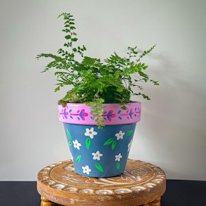 May include: A blue terracotta plant pot featuring hand-painted white flowers and green leaves, with a pink rim decorated with a magenta floral pattern. A green fern plant grows from the pot, which sits on a round, carved wooden pedestal.
