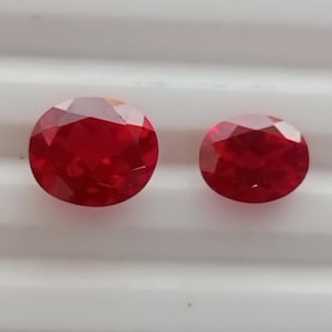 May include: Two faceted, oval-cut, deep red gemstones. The larger stone is on the left, with the smaller one to the right. The gems have a brilliant, reflective surface, suggesting high clarity and quality. The background is a white, ridged surface.