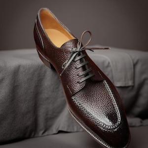 May include: A brown leather dress shoe with a textured finish and a classic lace-up design. The shoe features a rounded toe and a stitched sole. The laces are dark gray. The shoe is displayed on a gray surface.