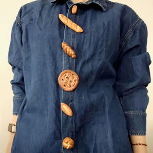 May include: A denim shirt with bread-shaped buttons. The buttons include a baguette, a round pie, and a pretzel. The shirt is a medium blue colour and has long sleeves.