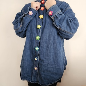 Peut inclure: Une chemise en jean bleu ornée de boutons en forme de fleurs multicolores. Les boutons sont rouges, roses, jaunes, verts et violets. La chemise a des manches longues et est boutonnée sur le devant.