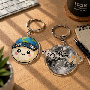 May include: Two acrylic keychains with silver key rings. One features a smiling moon wearing a blue cap with rockets and a world map. The other has a grayscale moon with the text "Copy Moon Joy."