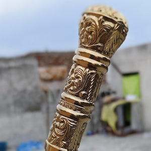 Może przedstawiać: Ozdobna laska do chodzenia ze szczegółowym, złotym metalowym uchwytem i ciemnobrązowym drewnianym trzonkiem. Uchwyt zdobią bogate wzory kwiatowe i zwoje, dodające elegancji.