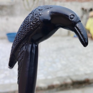 Puede incluir: Una cabeza de cuervo negra tallada corona un bastón. La cabeza del cuervo está detallada con plumas texturizadas y un pico curvo. El bastón es de un negro oscuro y brillante, lo que sugiere un acabado pulido. El fondo está borroso.