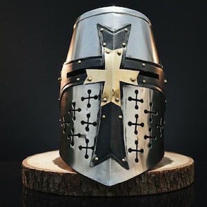 Puede incluir: Un casco medieval plateado y negro con una cruz dorada y detalles de remaches. El casco tiene cortes en forma de cruz y se exhibe sobre una base de madera. El diseño recuerda a una armadura histórica.