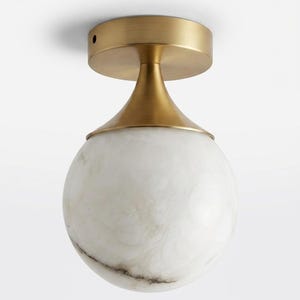 Op de afbeelding: Een plafondlamp met een bolvormige kap van wit marmer met grijze aders. De armatuur heeft een plafondkap en een conische hals van geborsteld messing die de ronde stenen bolkap verbindt met de cirkelvormige basis aan het plafond.