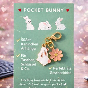 Könnte beinhalten: Ein Pocket Bunny-Anhänger mit einem weißen Hasen- und rosa Blumenanhänger. Der Anhänger ist an einem goldfarbenen Verschluss befestigt. Auf der Karte steht "Pocket Bunny" und der Text "Hier ist eine Umarmung, während ich nicht hier sein kann."