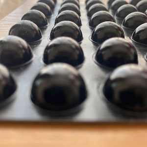 Puede incluir: Filas de objetos oscuros y brillantes en forma de cúpula dispuestos en una bandeja. Los objetos parecen ser de color oscuro, posiblemente chocolate o una confitería similar. La bandeja es de color gris claro y el fondo es de color marrón claro.