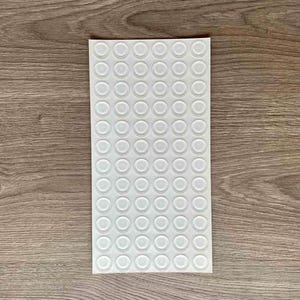 Può includere: Un foglio di cuscinetti adesivi trasparenti con un design circolare. I cuscinetti sono disposti a griglia su un foglio rettangolare bianco. Lo sfondo è una superficie in legno.