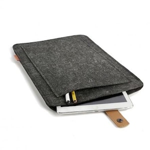 Pode incluir: Uma capa de tablet de feltro cinza escuro com fecho de tira de couro marrom. A capa tem um bolso contendo um tablet e duas canetas. O tablet é parcialmente visível. O fundo é branco.