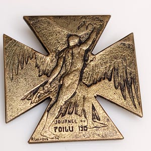 May include: A bronze-toned cross-shaped medal featuring a relief of a winged figure with outstretched arms. The text "JOURNEE DU POILU 1915" is embossed on the lower section, with the signature "R LALIQUE" visible in the bottom left corner.