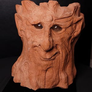 May include: A terracotta-colored sculpture of a tree-like face with a detailed, textured surface. The face has a prominent nose, a smiling mouth, and large, hollow eyes. The sculpture is likely a decorative item or art piece.