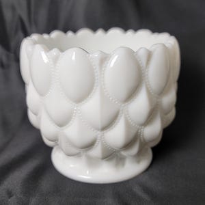 May include: A white, scalloped-rimmed bowl with an egg-shaped pattern. The bowl has a pedestal base and is made of opaque glass. The design features a series of raised, oval shapes with small dotted details, creating a textured surface.