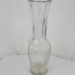 May include: A clear glass vase with a fluted design. The vase has a wide, open top and a slightly flared base. The glass is transparent, allowing a view of the interior. The vase is suitable for displaying flowers or as a decorative item.