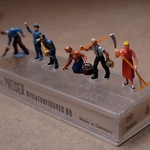 May include: Miniature HO scale figures of workers and a police officer. The figures are in various poses, including bending, carrying tools, and gesturing. The figures are on a clear plastic base with the text "ORIGINAL PREISER MINIATURFIGUREN HO" and "Made in Germany".