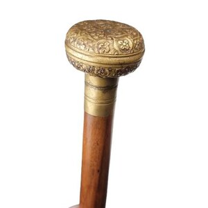 May include: A walking stick featuring a smooth, cylindrical wooden shaft and a rounded, ornate brass handle. The handle is decorated with intricate, embossed floral patterns and sits atop a plain, polished brass collar connecting to the wood.