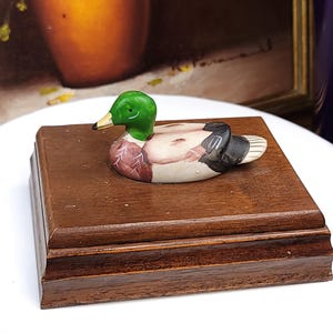 May include: A small ceramic mallard duck figurine with a vibrant green head, yellow beak, and brown and black body details, mounted securely on a rectangular, polished dark brown wooden base with a tiered edge design and smooth finish.