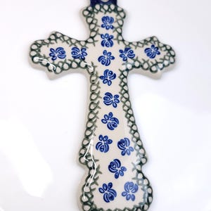 May include: A ceramic cross wall hanging featuring a cream-colored base with a hand-painted pattern of small blue flowers. The edges are decorated with a dark green cross-hatch design, and a dark blue loop is at the top for hanging purposes.