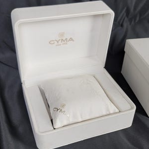 May include: A white watch box with the brand name "CYMA" printed on the inside lid and on a white pillow inside the box. A second white box is next to the open box. The boxes are on a dark surface.
