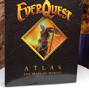 May include: A black, textured hardcover book titled EverQuest Atlas: The Maps of Myrist. The cover features a central diamond-shaped inset displaying a colorful illustration of a fantasy character holding a staff with a glowing light effect.