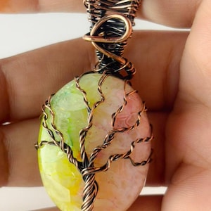 May include: A handcrafted pendant with a colourful, oval-shaped stone encased in copper wire. The stone shows shades of yellow, green, and pink. The wire is intricately woven to resemble a tree, creating a unique and artistic jewellery piece.