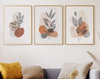 Puede incluir: Tres impresiones artísticas botánicas enmarcadas con marcos dorados cuelgan sobre un sofá gris con cojines decorativos. Dos mesas de centro doradas con tapas de mármol se asientan sobre una alfombra blanca y esponjosa. El arte presenta diseños de hojas abstractas.
