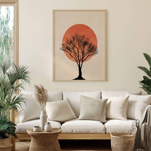 Puede incluir: Una impresión artística enmarcada que presenta una silueta de árbol minimalista contra un gran sol naranja. La obra de arte se exhibe sobre un sofá blanco con varias almohadas. La habitación tiene una paleta de colores neutros.