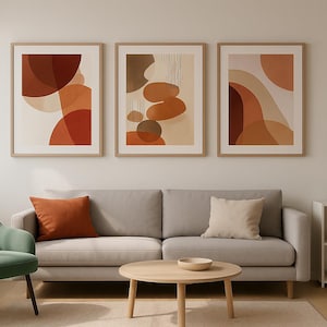 Puede incluir: Tres impresiones artísticas abstractas enmarcadas en tonos cálidos de naranja, marrón y beige cuelgan sobre un sofá gris. Un sillón verde, una mesa de centro de madera y una estantería blanca con una planta completan la escena.