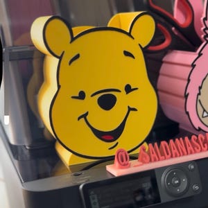May include: A yellow Winnie the Pooh-shaped desk organizer with black outlines. The organizer has a smiling face and is made of a solid material. The image also shows a pink object and a black printer.