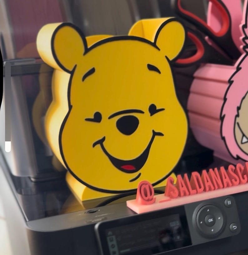 May include: A yellow Winnie the Pooh-shaped desk organizer with black outlines. The organizer has a smiling face and is made of a solid material. The image also shows a pink object and a black printer.