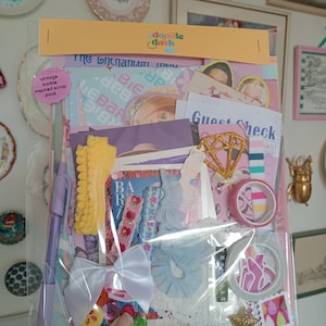 May include: A clear plastic bag containing scrapbooking materials. Inside are paper ephemera, ribbons, washi tape, and embellishments. The bag is labelled "doodle dash" and "vintage barbie inspired scrap pack". A pen and a white bow are visible.