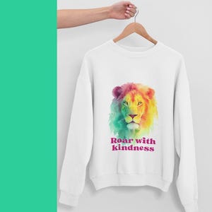 Può includere: Felpa bianca con un colorato disegno di leone ad acquerello. La criniera del leone è un mix di rosa, giallo e verde. Il testo "Roar with Kindness" è stampato sotto in rosa e viola.
