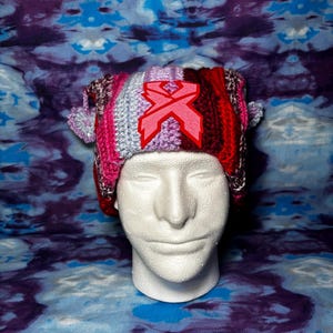 May include: A colorful, handmade crochet hat with a pink and red emblem. The hat features stripes of pink, red, purple, and gray. Two small pom-poms are attached to the sides. The hat is displayed on a white mannequin head.