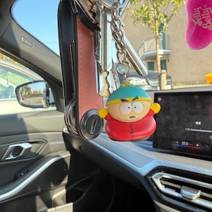 May include: A set of keys with a Yale key and a cartoon character keychain hanging inside a car. The keychain is a figure of a character wearing a red coat, yellow gloves, and a blue hat. The car interior is visible.