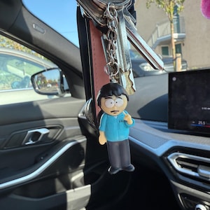 Puede incluir: Un juego de llaves con una etiqueta Yale y un llavero con un personaje de dibujos animados con una camisa azul y pantalones grises. El llavero cuelga dentro de un coche.
