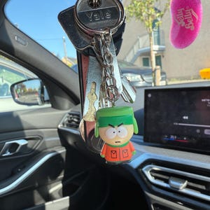 May include: A set of keys with a Yale key and a cartoon character keychain hanging in a car. The keychain is a small figure with a green hat, orange jacket, and red gloves. A pink Jelly Belly air freshener hangs in the background.
