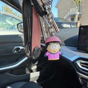 May include: A collection of keys with a pink leather key holder and a cartoon character keyring. The keyring is a figure wearing a pink hat and jacket. The keys are suspended inside a car.
