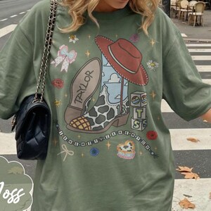 Puede incluir: Una camiseta verde musgo con un estampado gr&aacute;fico de una bota vaquera con patr&oacute;n de manchas de vaca, un sombrero rojo, el nombre Taylor, el n&uacute;mero 13 y bloques con las letras T, S. El dise&ntilde;o incluye una cadena de cuentas con el texto THE NEW WITTY TEN OR YOU.