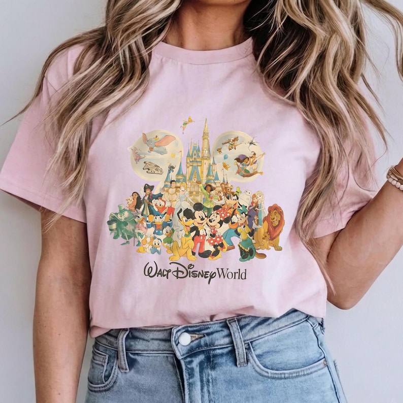 Puede incluir: Una camiseta de manga corta de color rosa claro con un estampado gr&aacute;fico colorido de varios personajes de Disney y la silueta de un castillo en forma de cabeza de Mickey Mouse, con el texto Walt Disney World impreso en negro debajo.