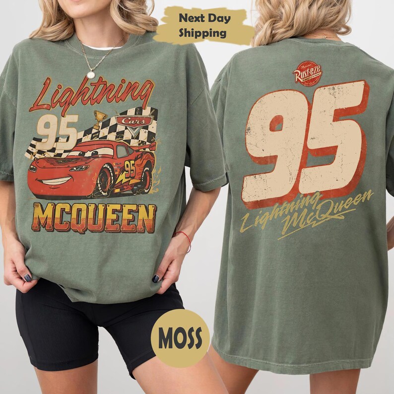 May include: An olive green t-shirt featuring a graphic of the red race car Lightning McQueen with the number 95 and a checkered flag on the front. The back of the shirt displays a large number 95 in beige with red outlines and the text Lightning McQueen.
