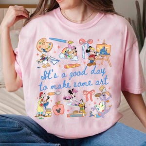 May include: A light pink t-shirt featuring a graphic print of various Disney characters engaged in artistic activities, including painting and drawing. The text "It's a good day to make some art" is printed in blue in the center of the shirt design.