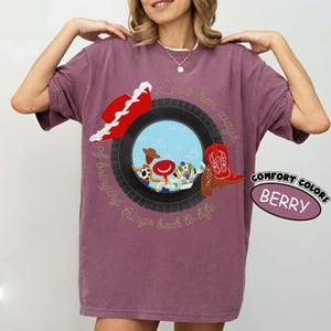May include: A berry-colored t-shirt featuring a graphic of a black tire framing cartoon characters Woody, Jessie, and Buzz Lightyear. The design includes a red cowboy hat, a red cowboy boot, and the text Love has ways of bringing things back to life.