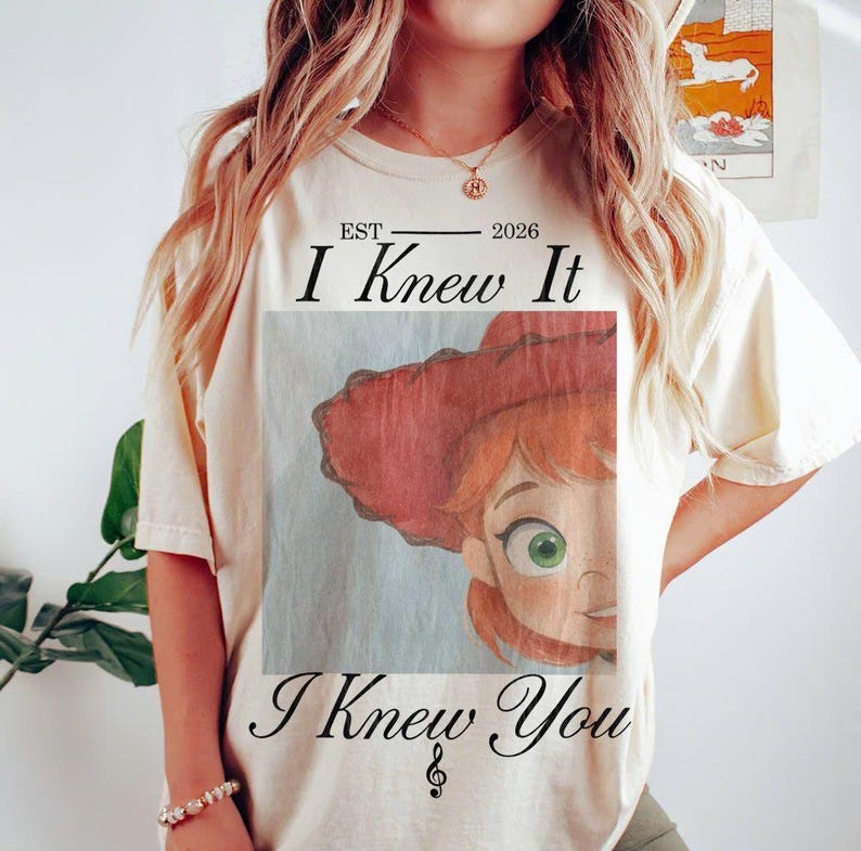 May include: A cream-colored short-sleeve t-shirt featuring a graphic of a red-hatted character with green eyes. The text reads EST 2026 I Knew It I Knew You with a small musical note symbol below the text, printed in a black serif font.