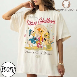 May include: Ivory t-shirt with a graphic of Disney's "The Three Caballeros." The design features Donald Duck, Panchito Pistoles, and Jos&eacute; Carioca. The shirt has the text "Walt Disney Presents The Three Caballeros" and "Souvenir Album."
