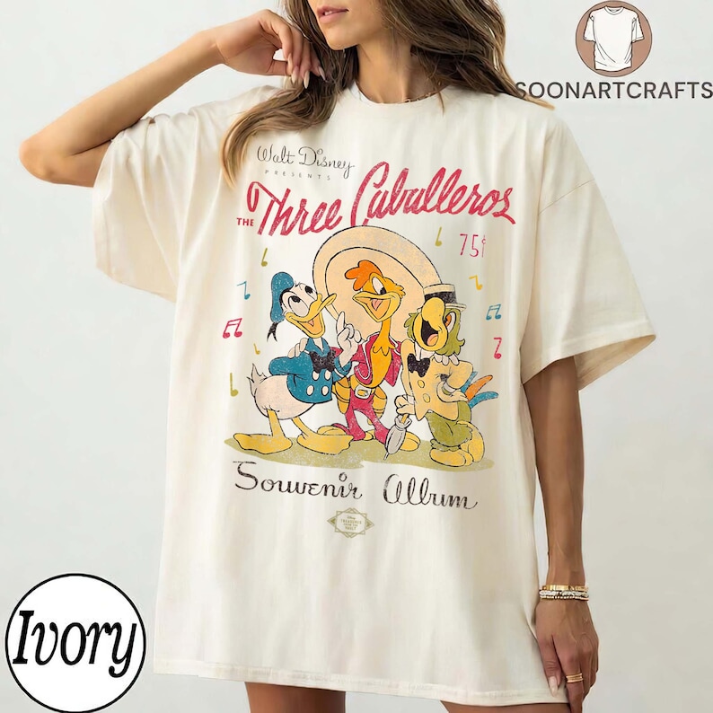 May include: Ivory t-shirt with a graphic of Disney's "The Three Caballeros." The design features Donald Duck, Panchito Pistoles, and Jos&eacute; Carioca. The shirt has the text "Walt Disney Presents The Three Caballeros" and "Souvenir Album."