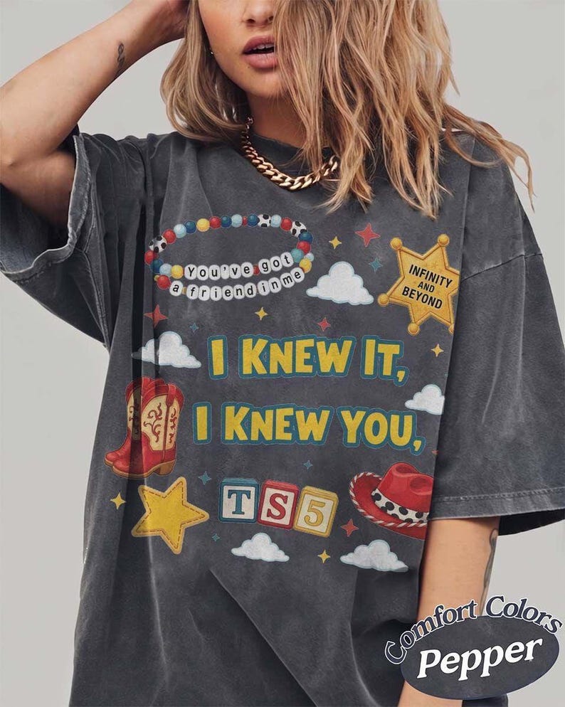 May include: A charcoal gray short-sleeve t-shirt featuring a graphic print with the text I KNEW IT, I KNEW YOU, TS5, INFINITY AND BEYOND, and YOU'VE GOT A FRIEND IN ME. The design includes a beaded bracelet, a yellow sheriff star, clouds, a red cowboy boot, and a red cowboy hat.