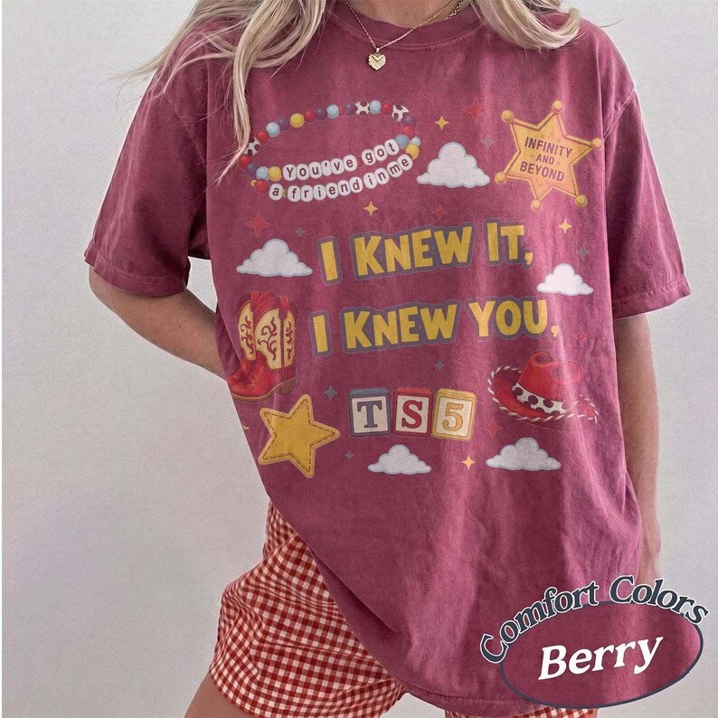 May include: A berry-colored short-sleeve t-shirt featuring a graphic print with the text "You've got a friend in me," "I KNEW IT, I KNEW YOU," and "TS5." The design includes a beaded necklace, a yellow star, a red cowboy boot, and a hat.